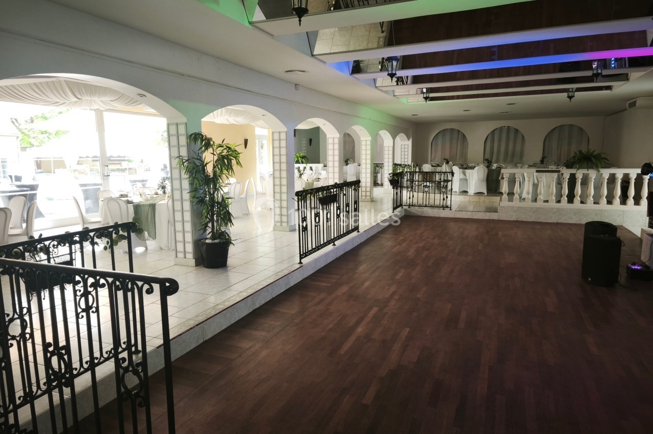 Salle de réception lumineuse avec parquet, tables dressées, plantes décoratives et balustrades en fer forgé.