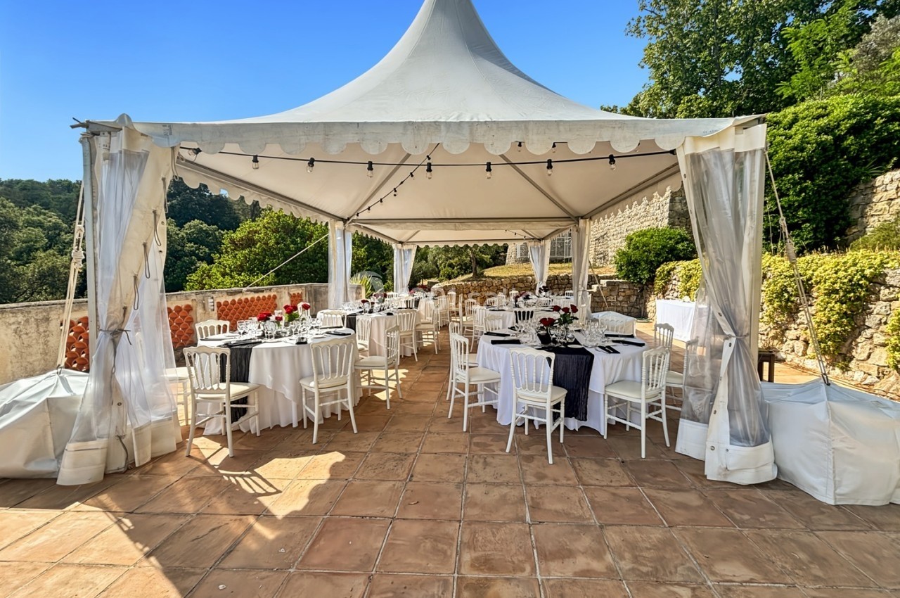 Tente blanche aménagée avec des tables et chaises pour un événement en extérieur, entourée de verdure et de murs en pierre.