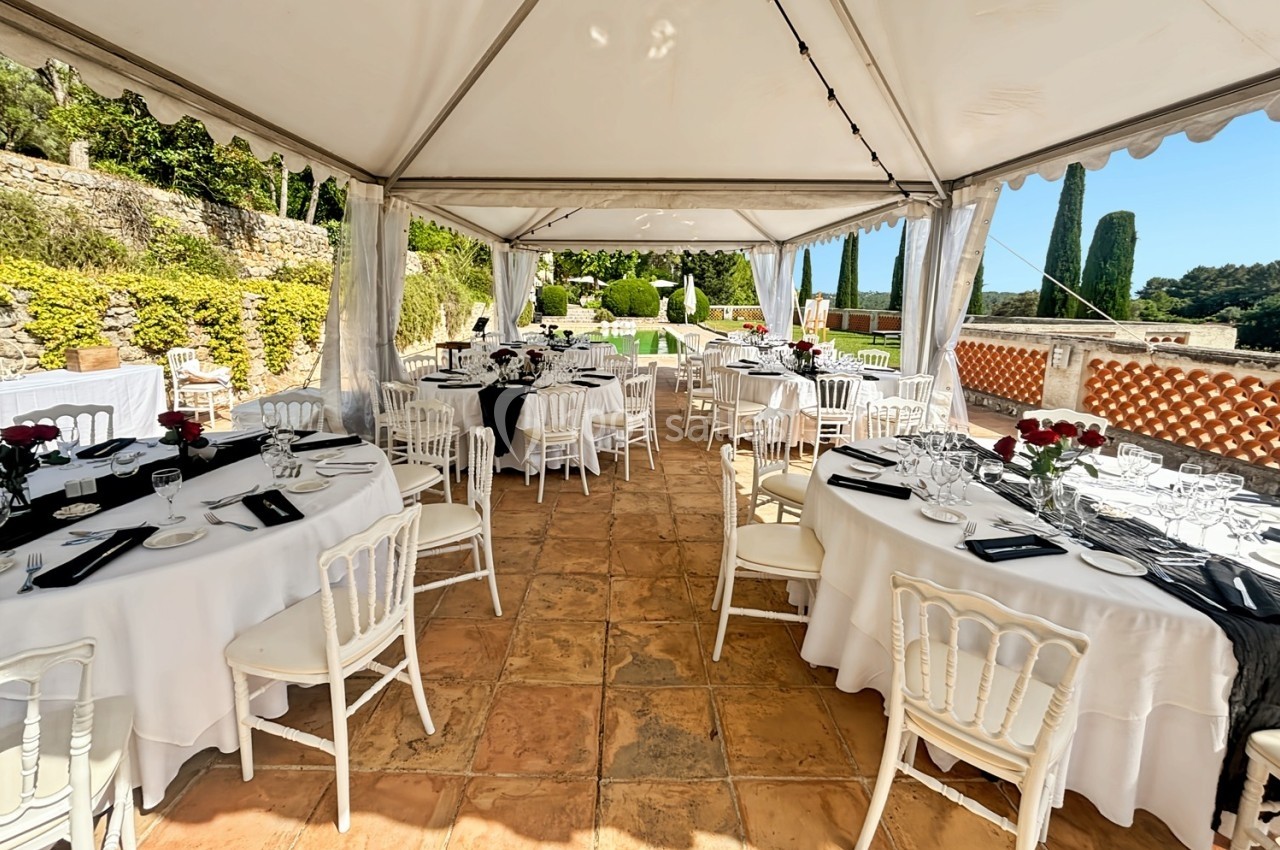 Tables rondes dressées sous une tente blanche sur une terrasse ensoleillée, entourée de verdure et de murs en pierre.