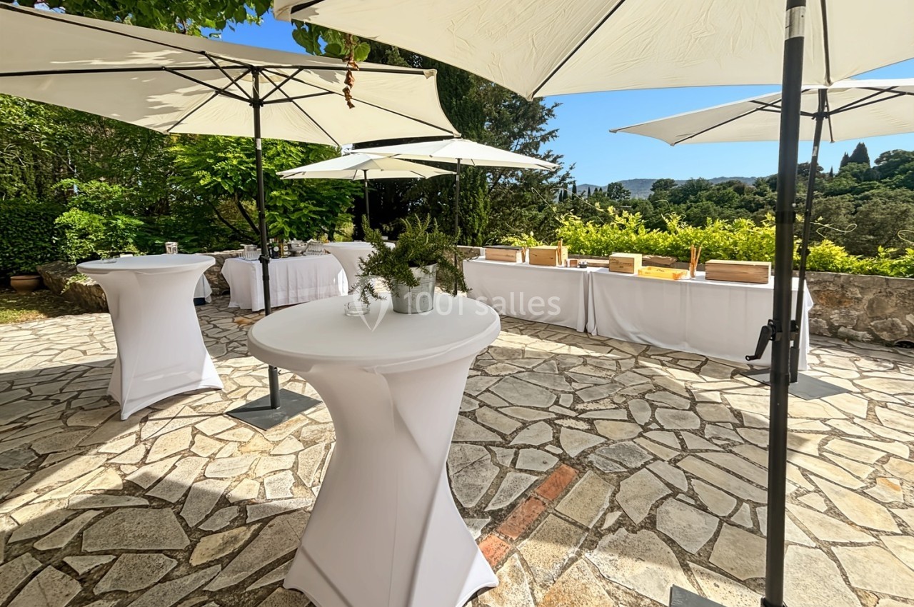Terrasse ensoleillée avec tables hautes et parasols blancs, entourée de verdure et vue sur un paysage arboré.
