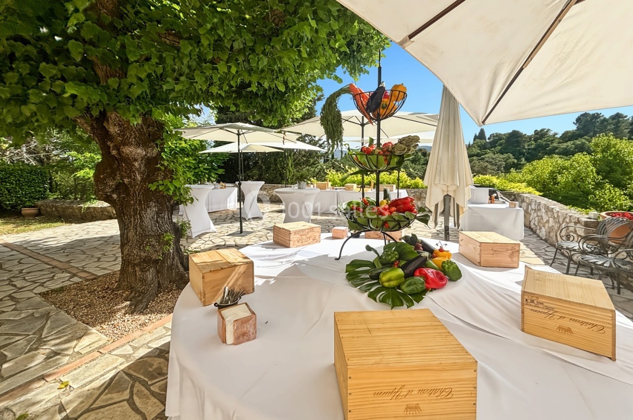 Table décorée en extérieur avec fruits, légumes, caisses en bois et parasols, entourée de verdure et d'arbres.