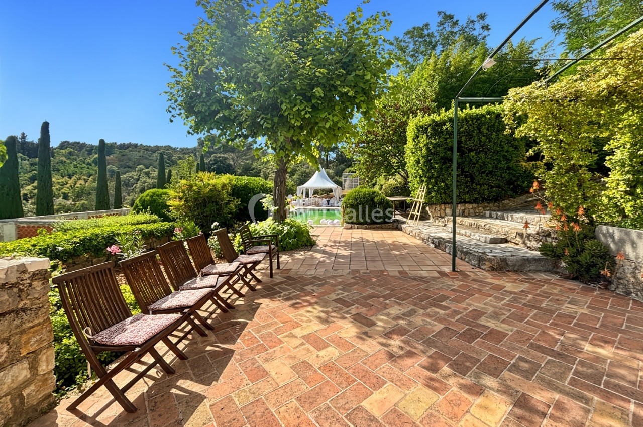 Terrasse en briques avec chaises en bois, entourée de verdure et d'arbres, sous un ciel bleu dégagé.