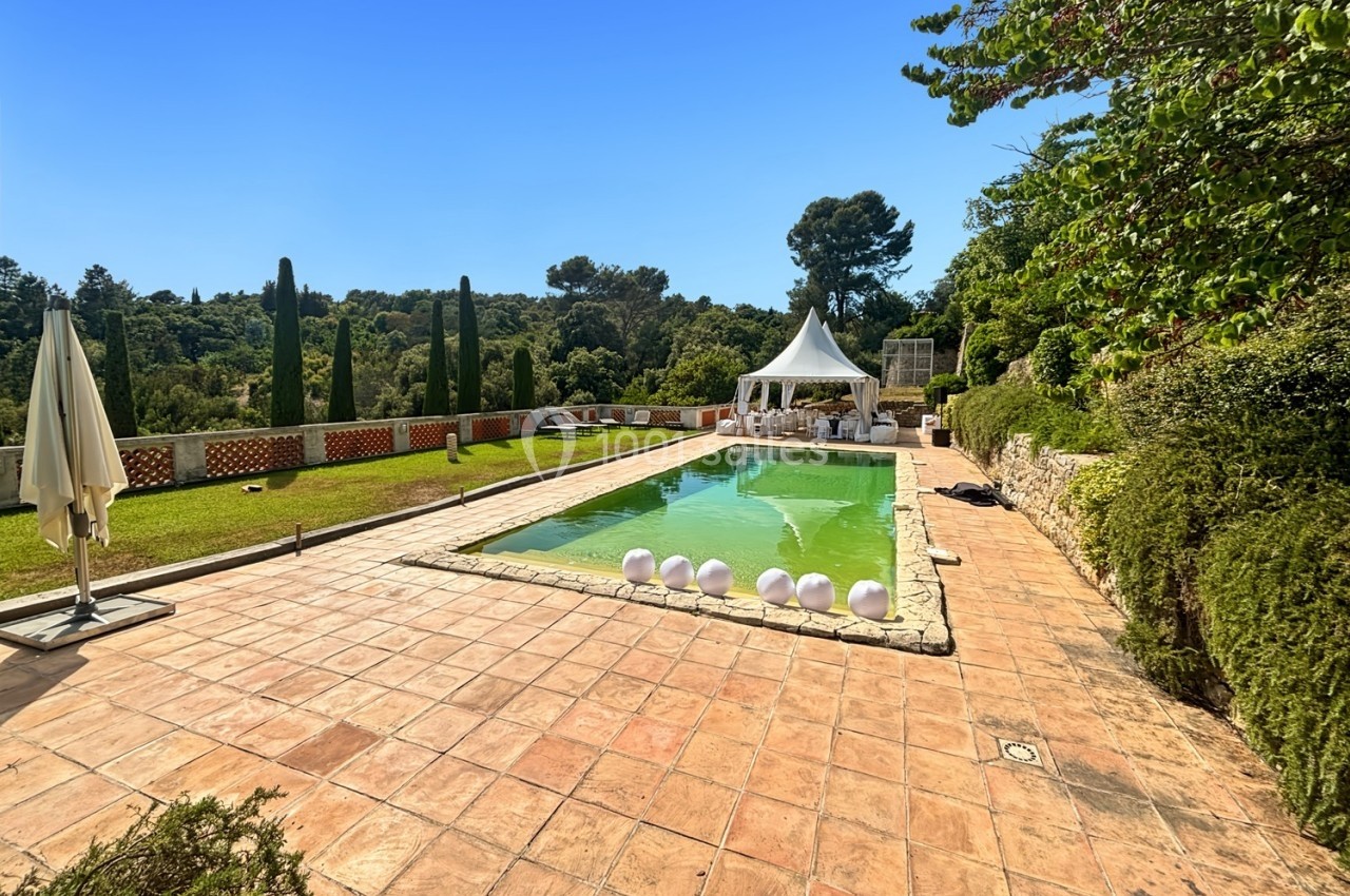 Piscine extérieure entourée de végétation, avec terrasse en pierre, transats et un chapiteau blanc en arrière-plan.