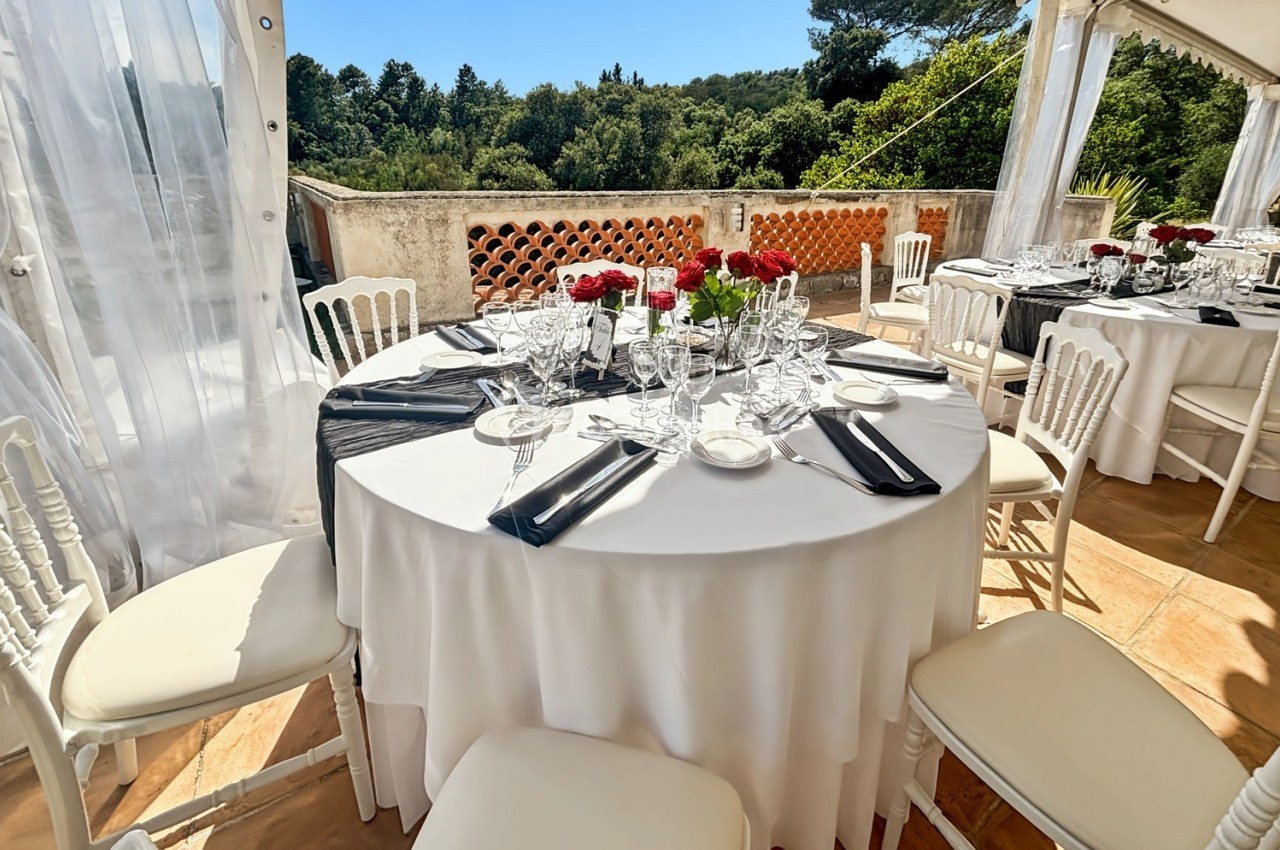 Table dressée en extérieur sous une tente, avec nappes blanches, serviettes noires et bouquets de roses rouges.