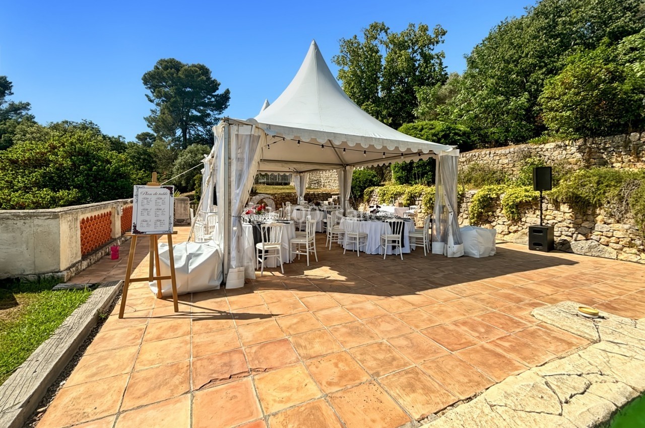 Tente blanche installée sur une terrasse ensoleillée avec des tables et chaises, entourée de verdure et de murs en pierre.