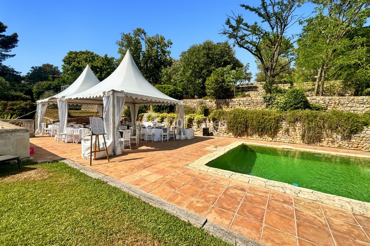 Tente blanche aménagée avec tables et chaises près d'une piscine entourée de végétation et de murs en pierre.