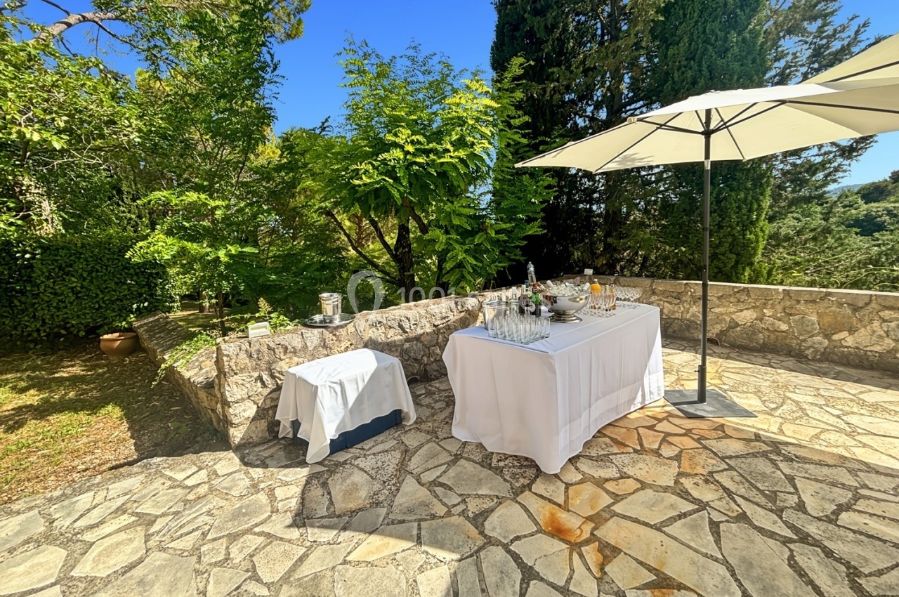 Table dressée avec boissons et vaisselle sous un parasol sur une terrasse en pierre entourée de végétation.