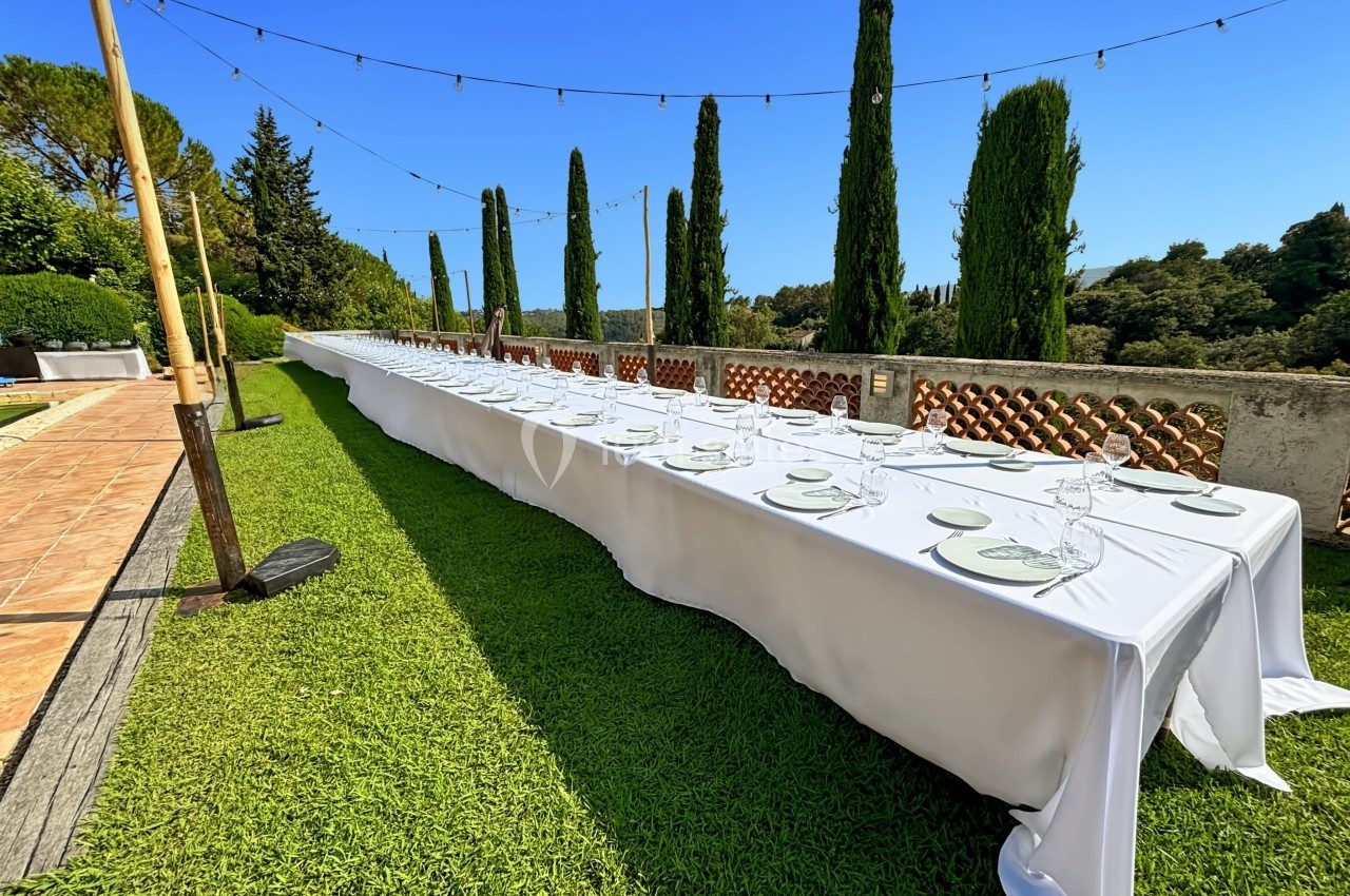 Grande table extérieure dressée avec nappes blanches, assiettes et verres, dans un jardin bordé de cyprès.