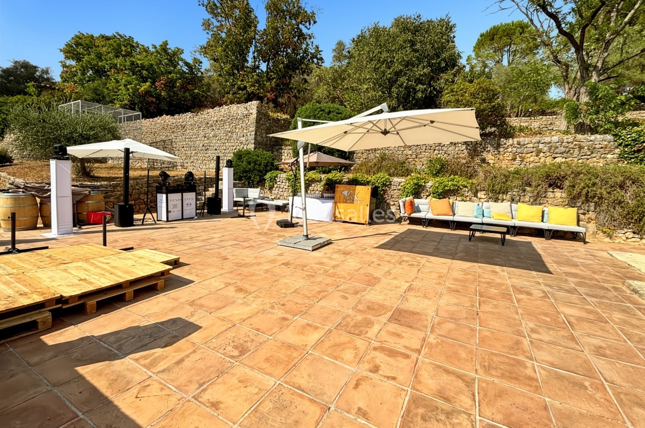 Terrasse ensoleillée avec sol en dalles, parasols, bancs colorés et végétation en arrière-plan.