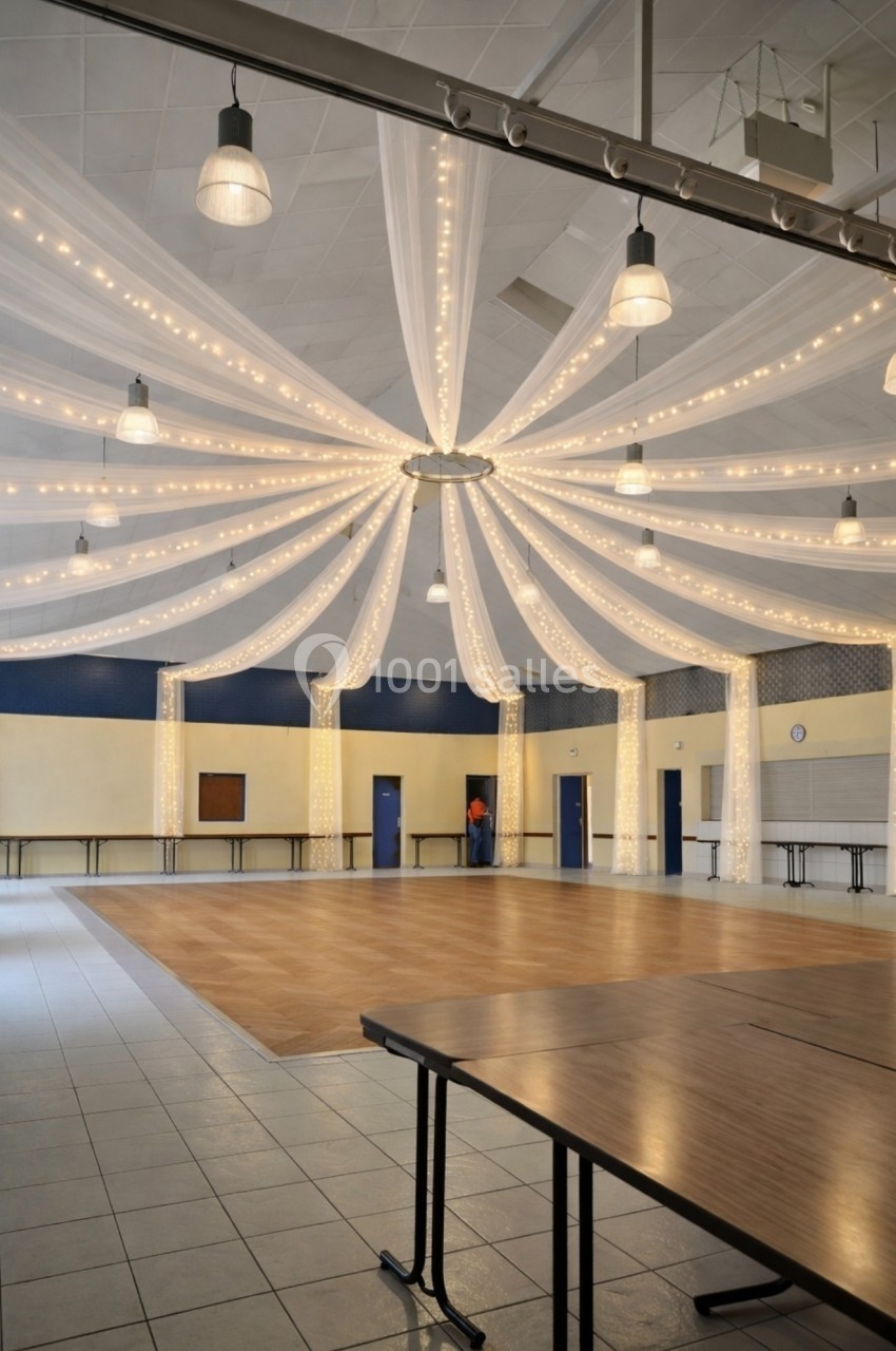 Salle de réception avec plafond décoré de voilages et guirlandes lumineuses, tables pliées au premier plan.