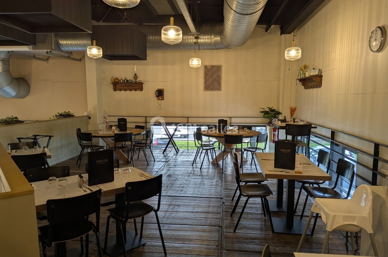 Salle de restaurant avec tables dressées, chaises noires et décoration sobre, éclairée par des suspensions modernes.
