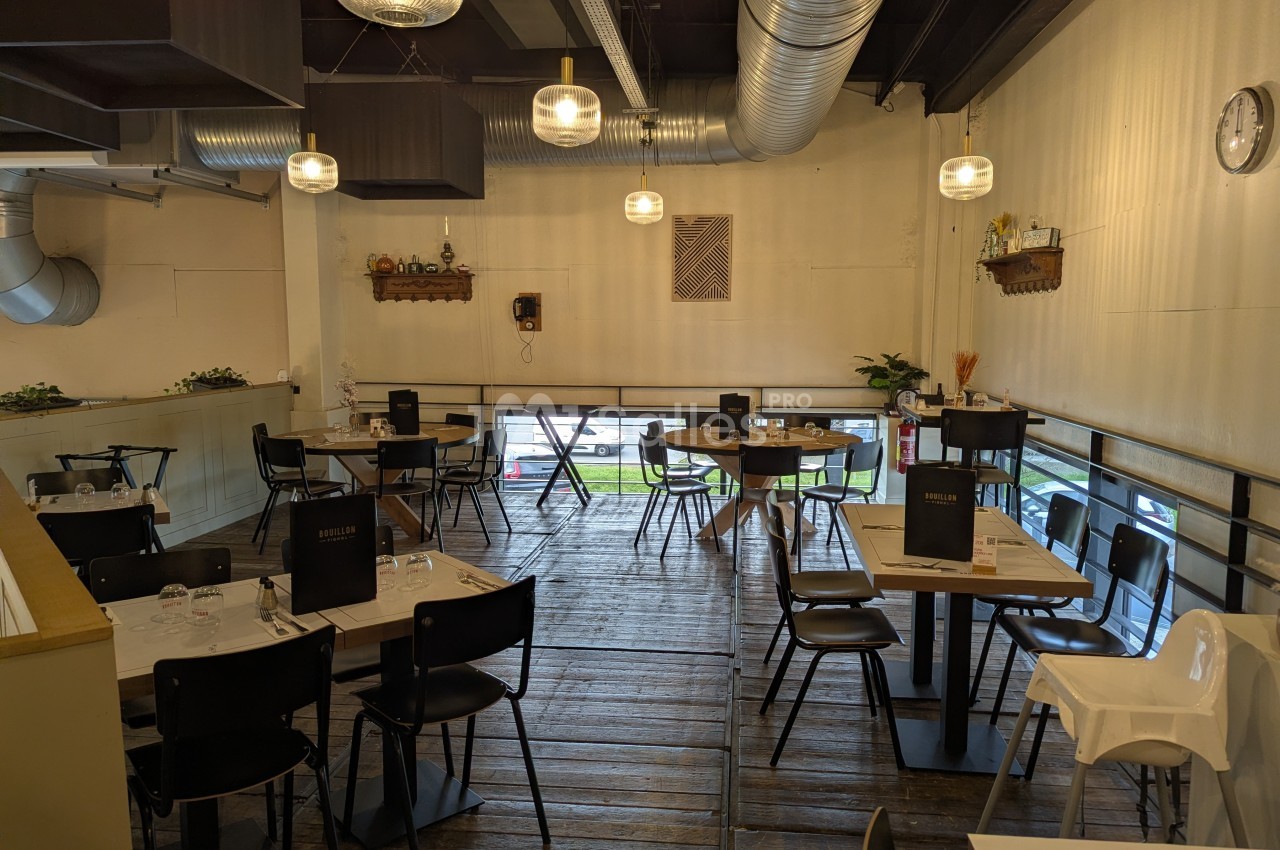 Salle de restaurant avec tables dressées, chaises noires et décoration sobre, éclairée par des suspensions modernes.