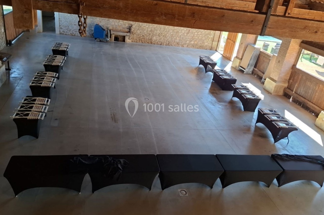 Salle spacieuse avec tables noires alignées et chaises pliées, dans un espace en pierre avec poutres apparentes.