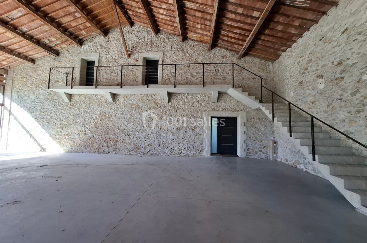 Intérieur d'un bâtiment avec murs en pierre, plafond en bois, mezzanine avec garde-corps et escalier en béton.