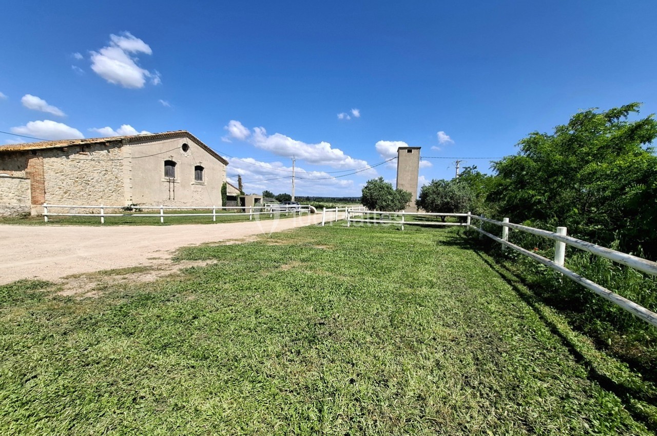 Cour herbeuse bordée de clôtures blanches, avec un bâtiment en pierre et une tour sous un ciel bleu parsemé de nuages.