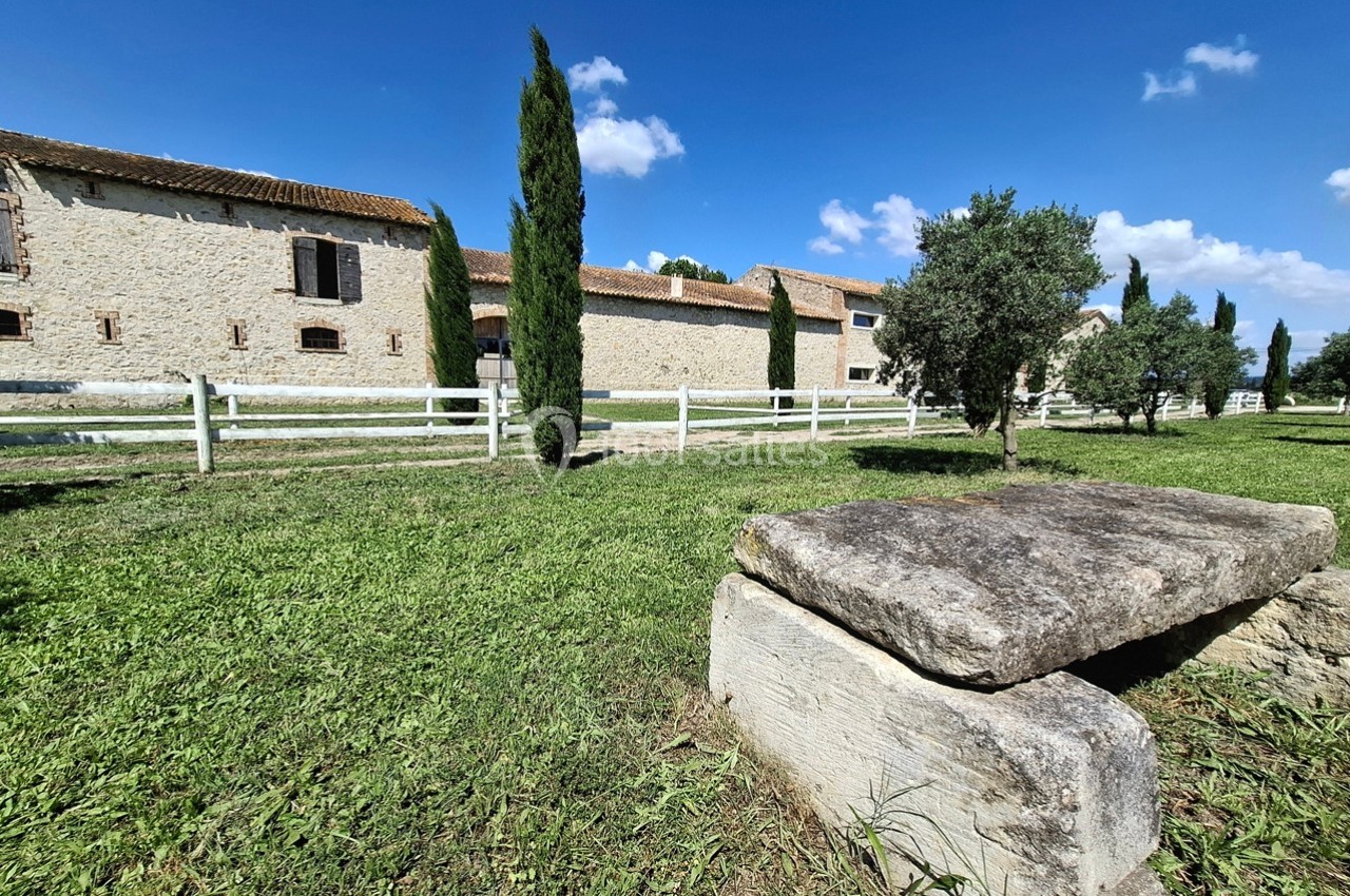 Bâtiment en pierre avec toit en tuiles, entouré de cyprès, d'oliviers et d'une clôture blanche, sous un ciel bleu.