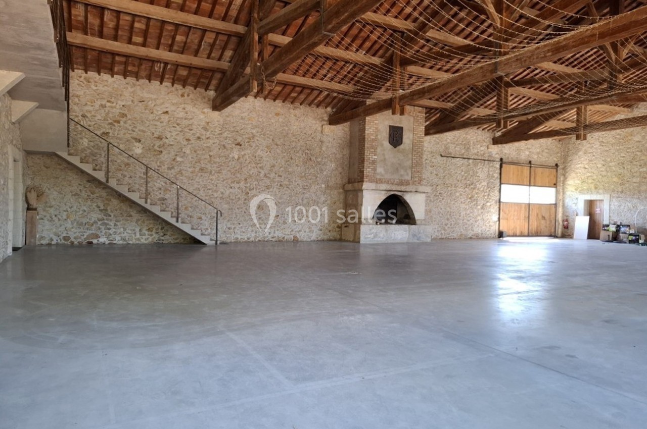 Intérieur d'une grande salle en pierre avec un sol en béton, une cheminée, une mezzanine et une porte en bois.