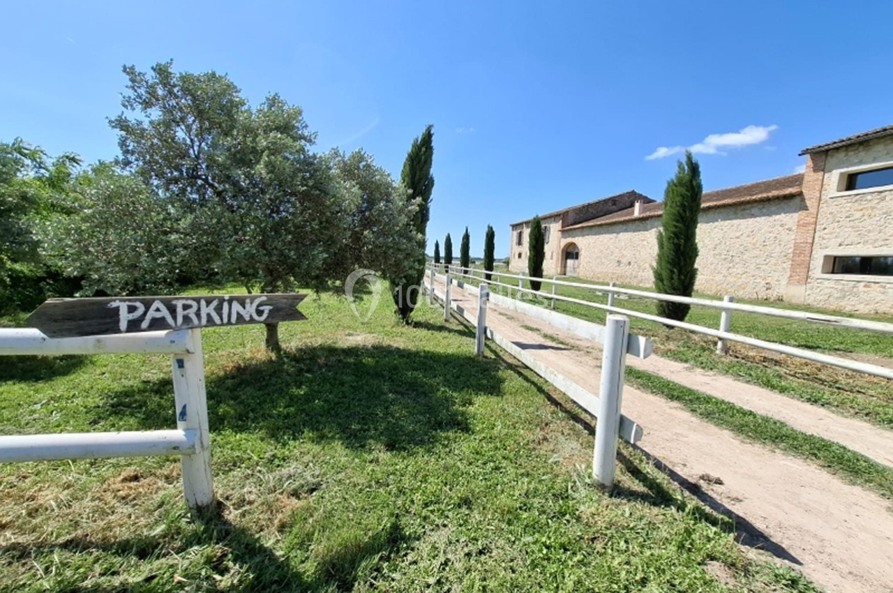 Chemin en terre bordé de barrières blanches menant à un bâtiment en pierre, avec un panneau indiquant ’Parking’.