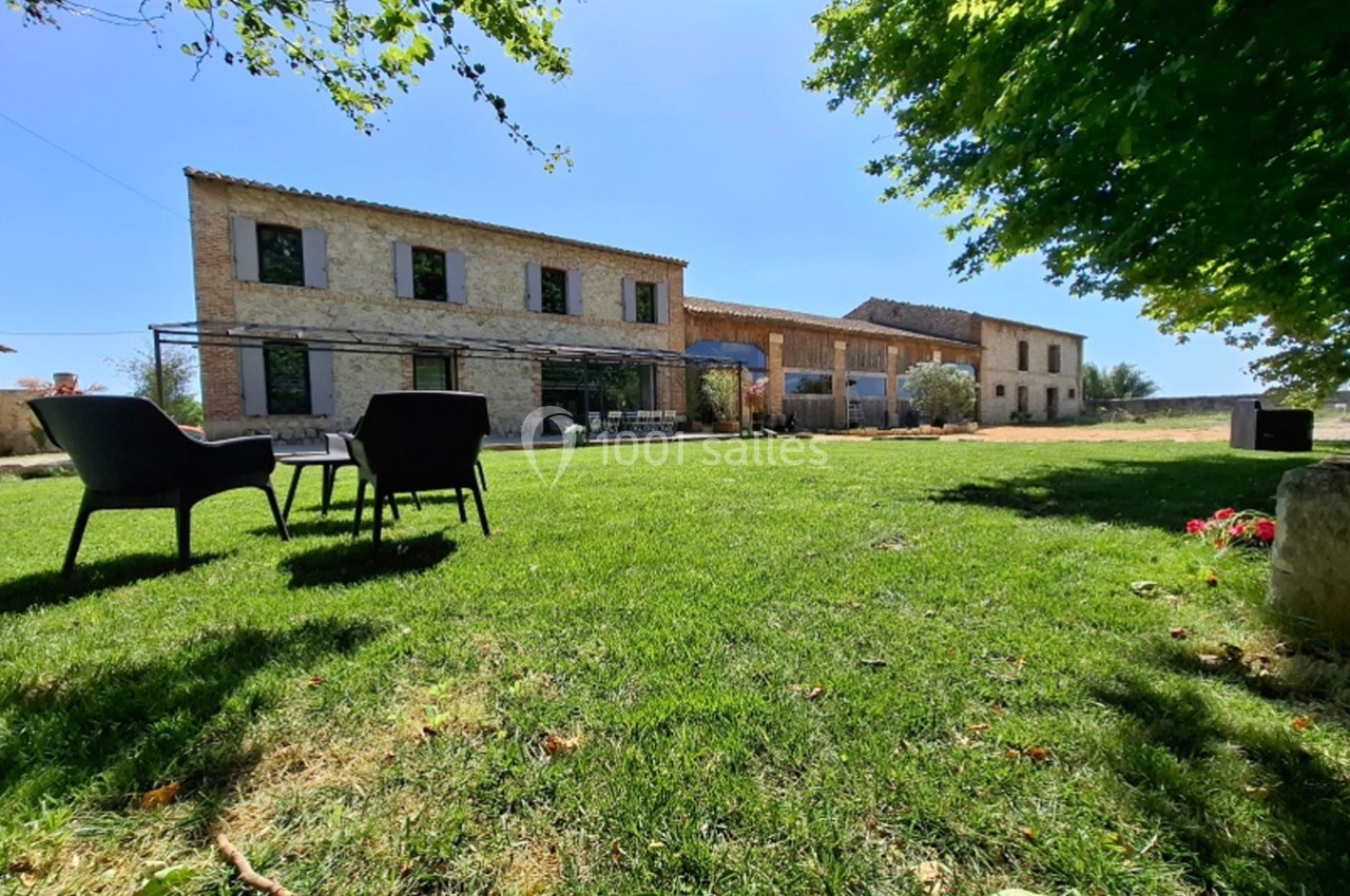 Grande maison en pierre avec jardin verdoyant, deux chaises noires au premier plan sous un arbre.