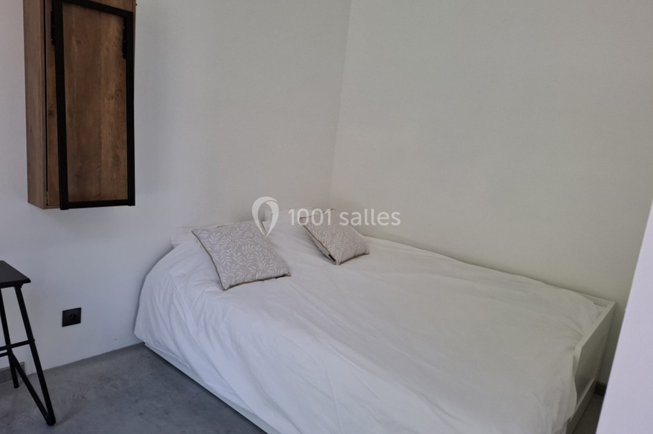Lit double avec draps blancs et deux coussins décoratifs, dans une chambre minimaliste aux murs blancs.