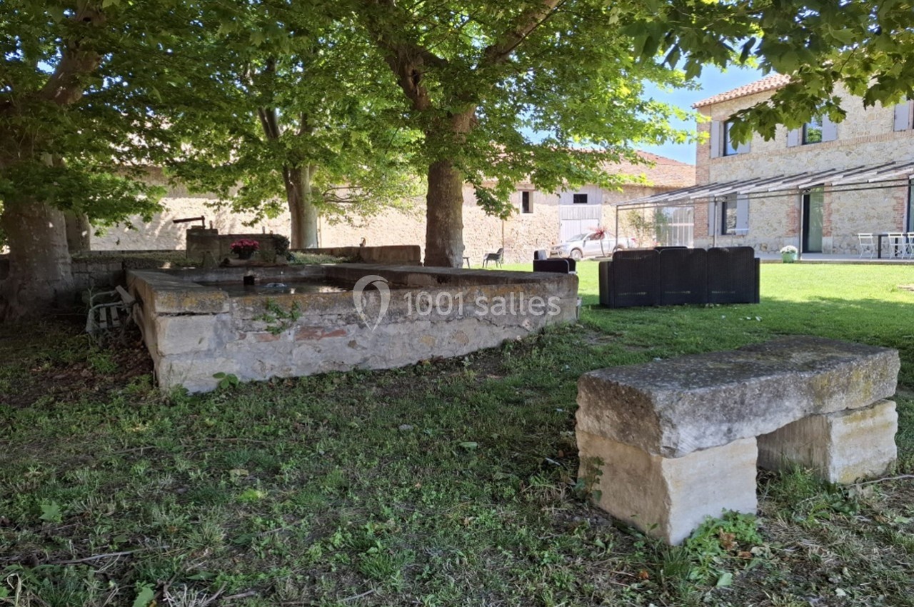 Ancien lavoir en pierre entouré d'arbres, avec des bâtiments en pierre et du mobilier de jardin en arrière-plan.