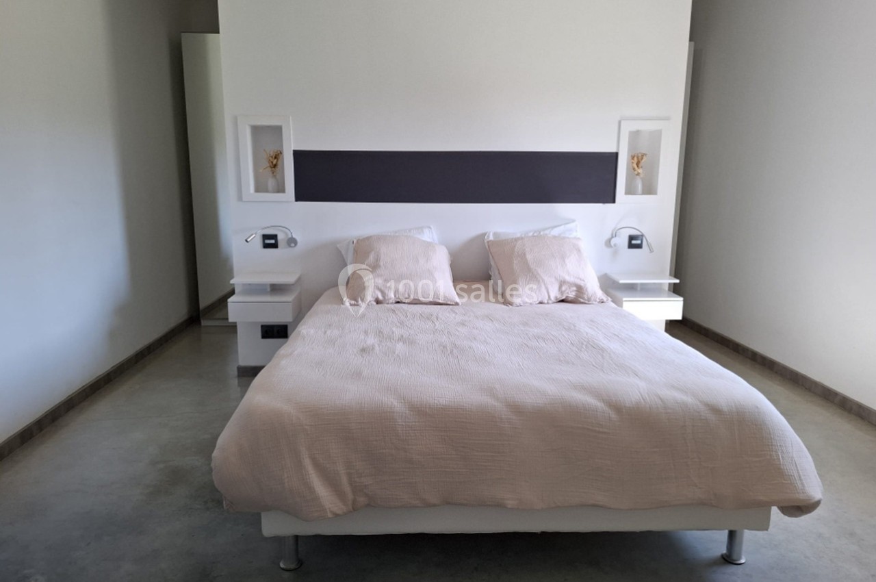 Lit double avec draps beiges, encadré par deux tables de chevet blanches intégrées à une tête de lit moderne.