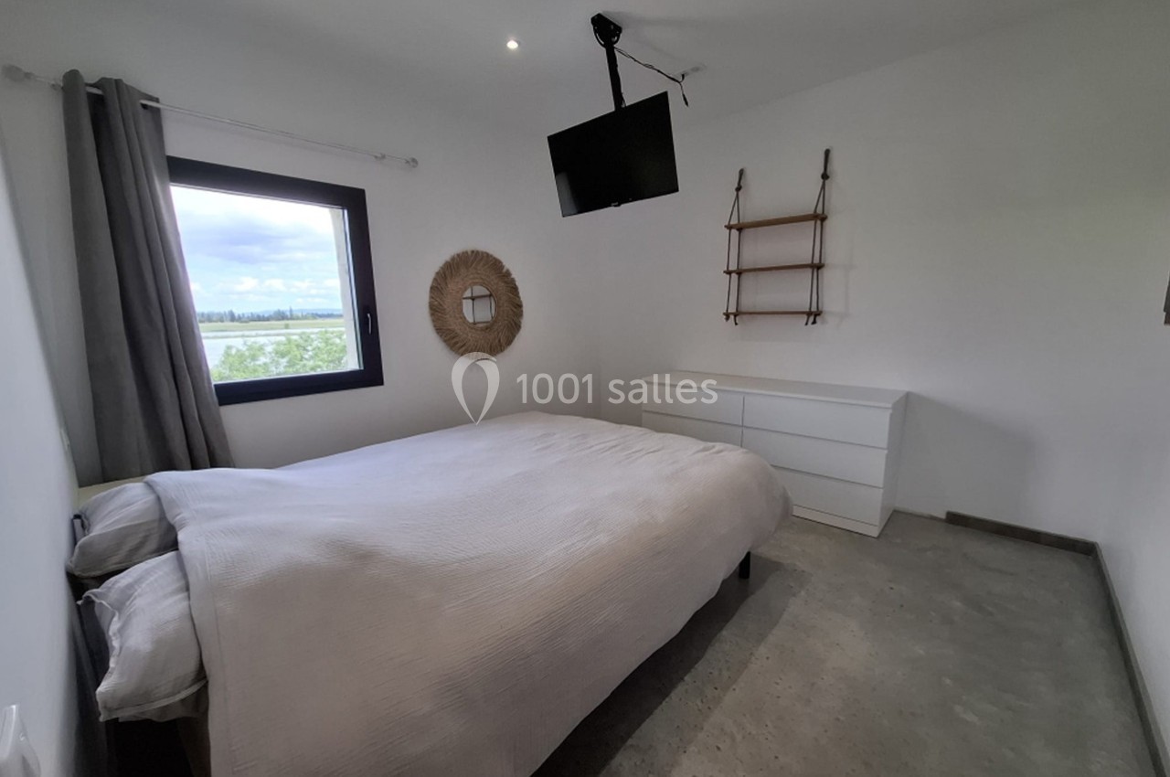 Chambre lumineuse avec lit double, commode blanche, étagères murales, miroir rond et fenêtre donnant sur un paysage.