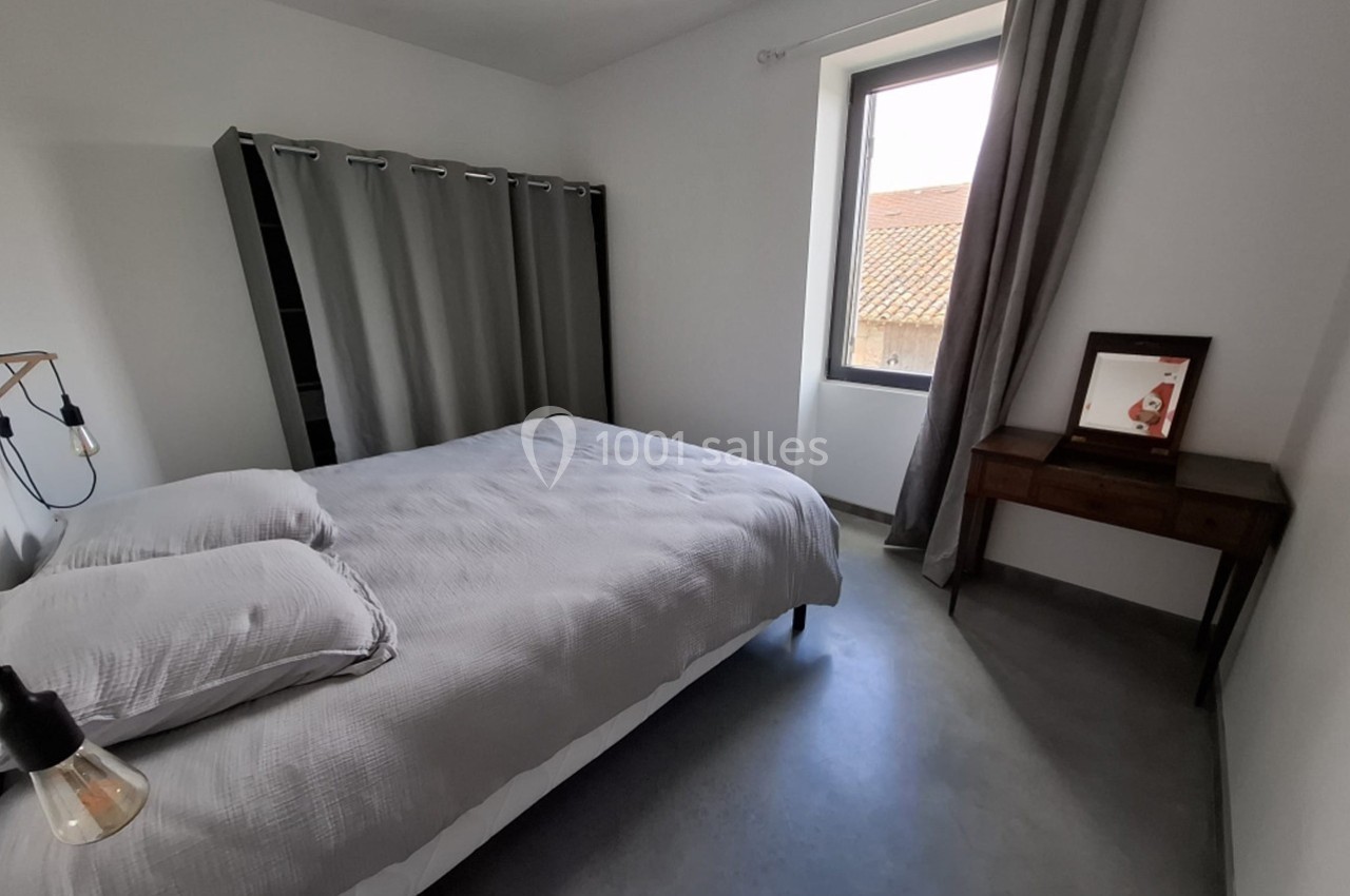 Chambre lumineuse avec lit double, rideaux gris, bureau en bois et fenêtre donnant sur un toit en tuiles.