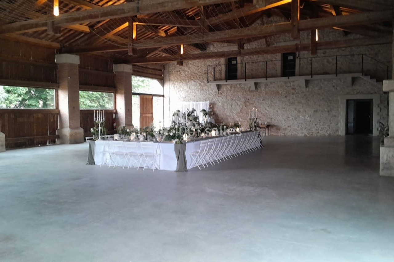 Grande salle en pierre avec une longue table décorée pour un événement, éclairée par une lumière naturelle et des lampes.