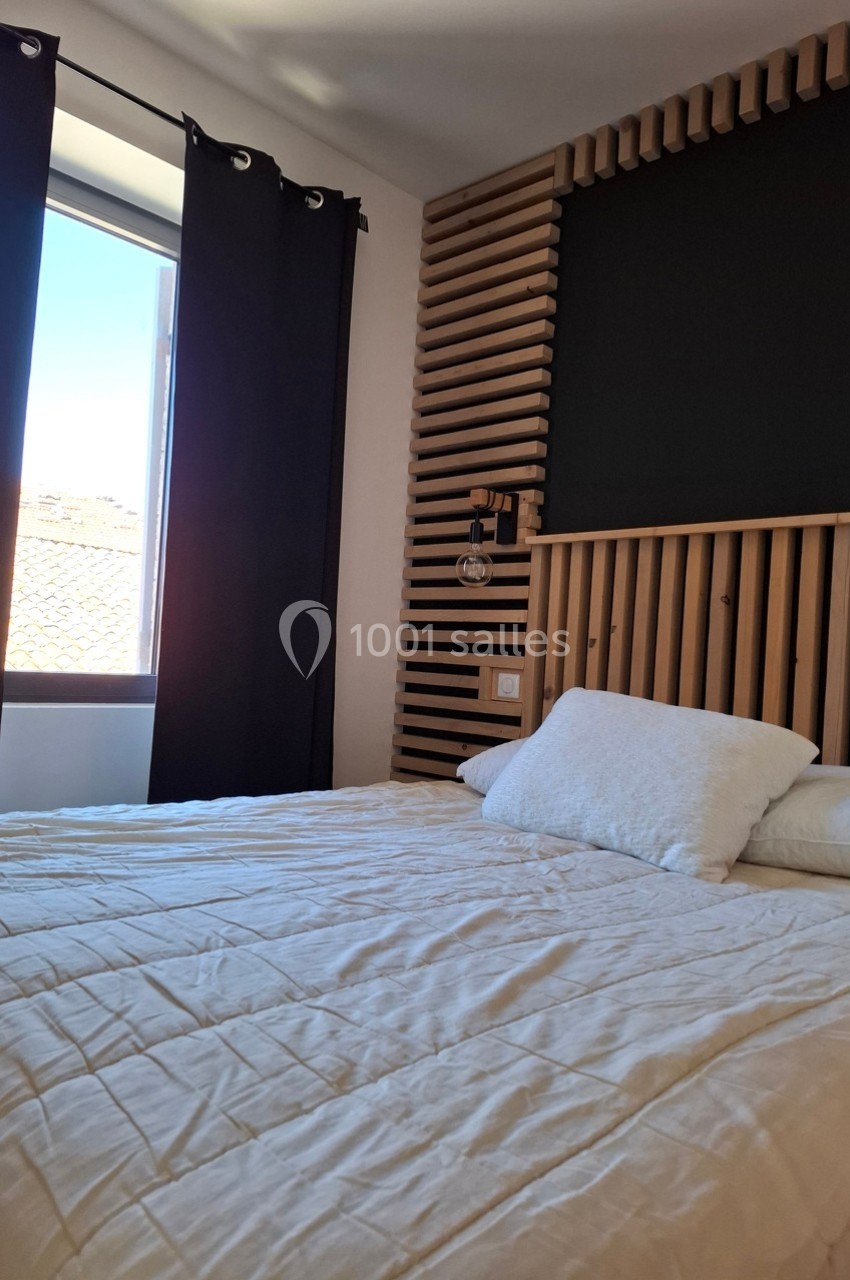 Lit double avec draps blancs dans une chambre lumineuse, avec une fenêtre ouverte et un mur en bois décoratif.