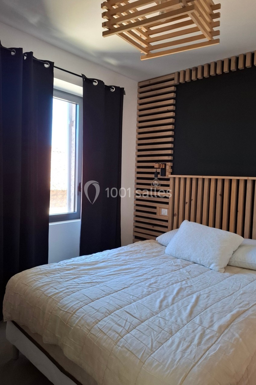 Chambre avec lit double, tête de lit en bois, rideaux noirs et fenêtre laissant entrer la lumière naturelle.