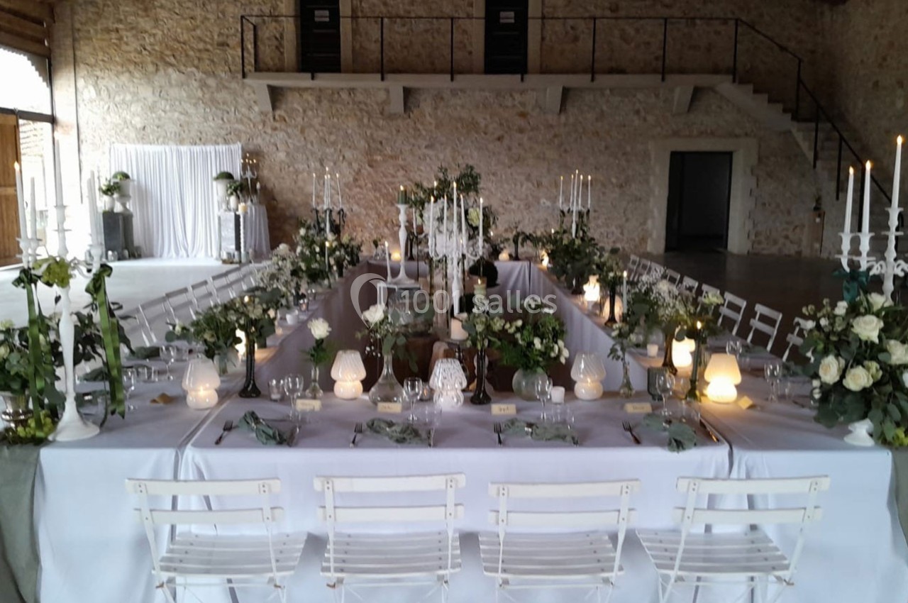 Grande table rectangulaire décorée de fleurs blanches et bougies, installée dans une salle en pierre avec un escalier au…