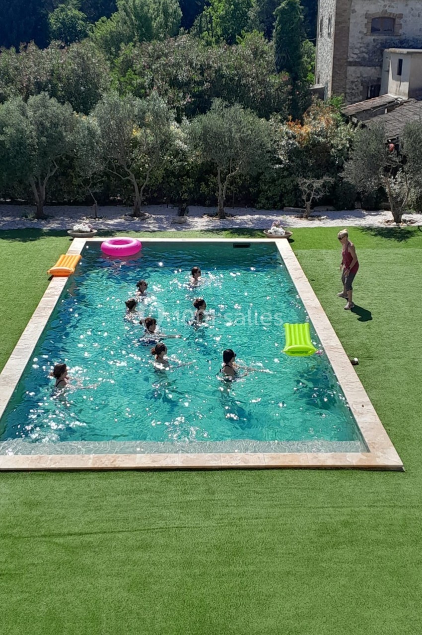 Des personnes nagent et jouent dans une piscine extérieure entourée de pelouse et d'arbres.