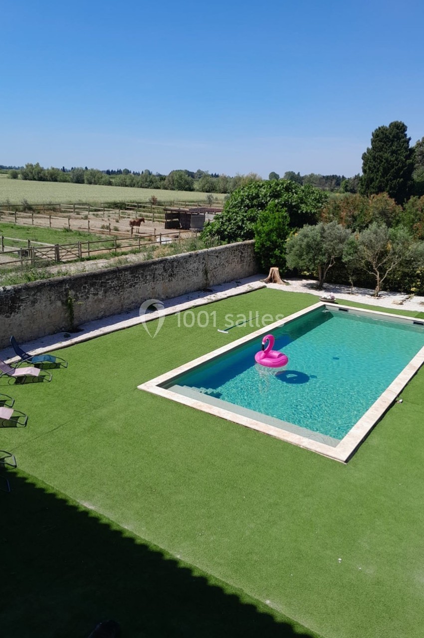 Piscine extérieure entourée de pelouse artificielle, avec une bouée rose en forme de flamant rose, près d'un champ.
