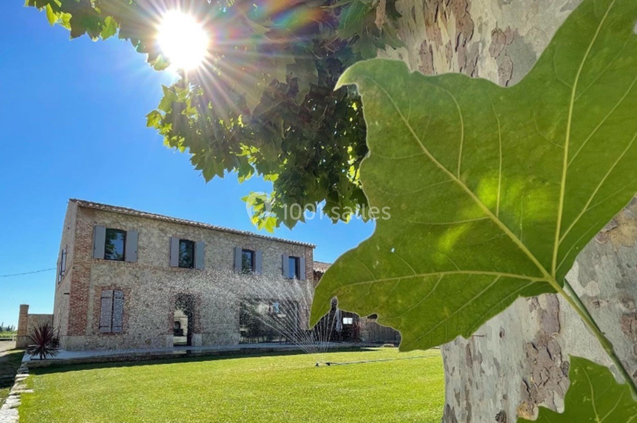 Maison en pierre avec volets verts, pelouse arrosée, arbre au premier plan et soleil éclatant dans un ciel bleu.