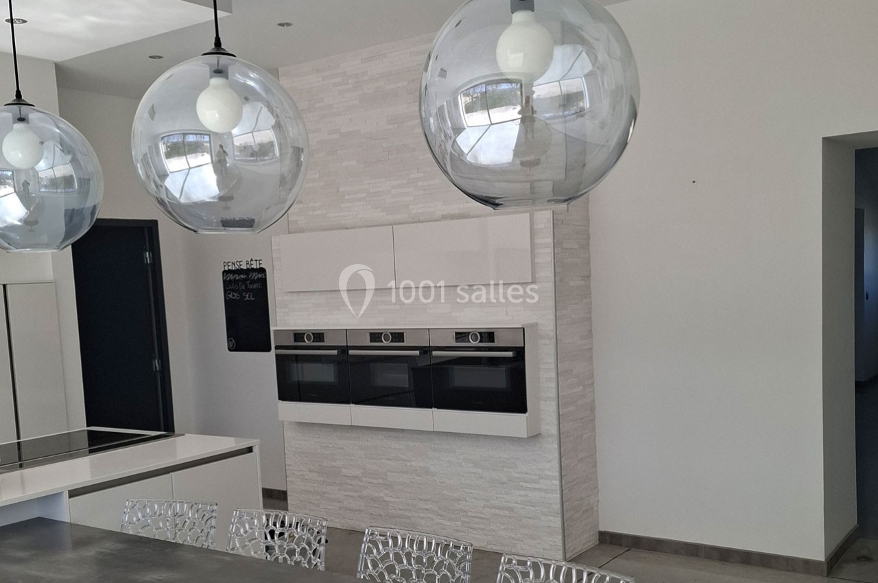 Cuisine moderne avec îlot central, luminaires suspendus sphériques et mur équipé de fours encastrés.