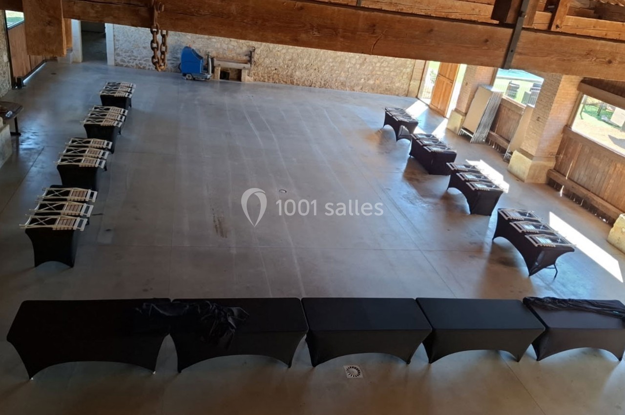 Salle vide avec des tables noires alignées en U, des chaises pliées et un sol en béton dans un espace ouvert avec poutres en…