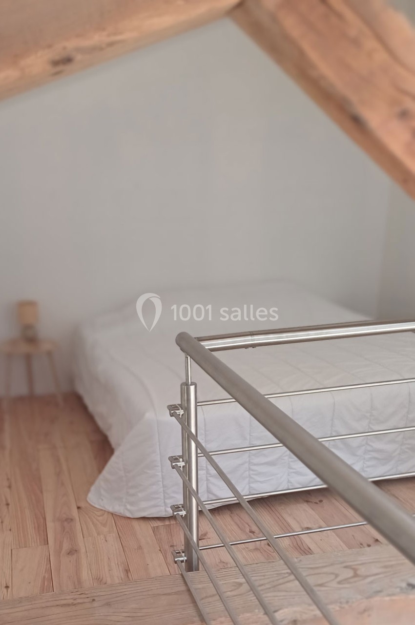 Lit double avec drap blanc dans une chambre mansardée en bois, vue depuis une rambarde métallique.