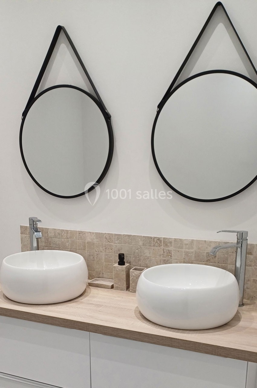 Deux lavabos ronds blancs sur un plan en bois clair, avec deux miroirs ronds suspendus par des sangles noires.