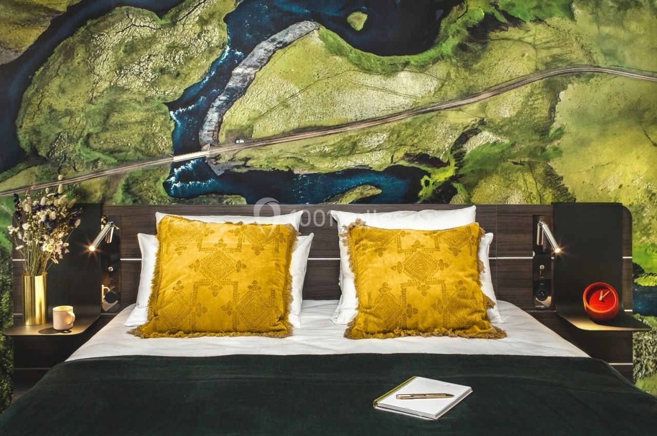Lit double avec coussins jaunes, couverture verte et décoration murale représentant une vue aérienne de paysage naturel.