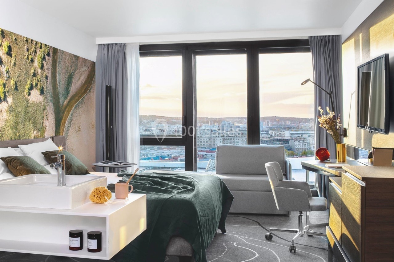 Chambre d'hôtel moderne avec lit, bureau, fauteuil, grande fenêtre offrant une vue sur la ville au coucher du soleil.