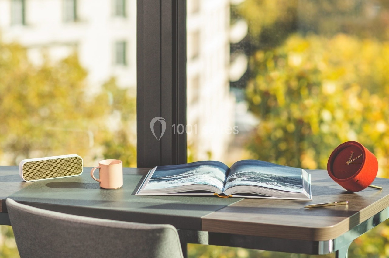 Livre ouvert sur un bureau près d'une fenêtre, entouré d'une tasse, d'une horloge rouge et d'une enceinte.