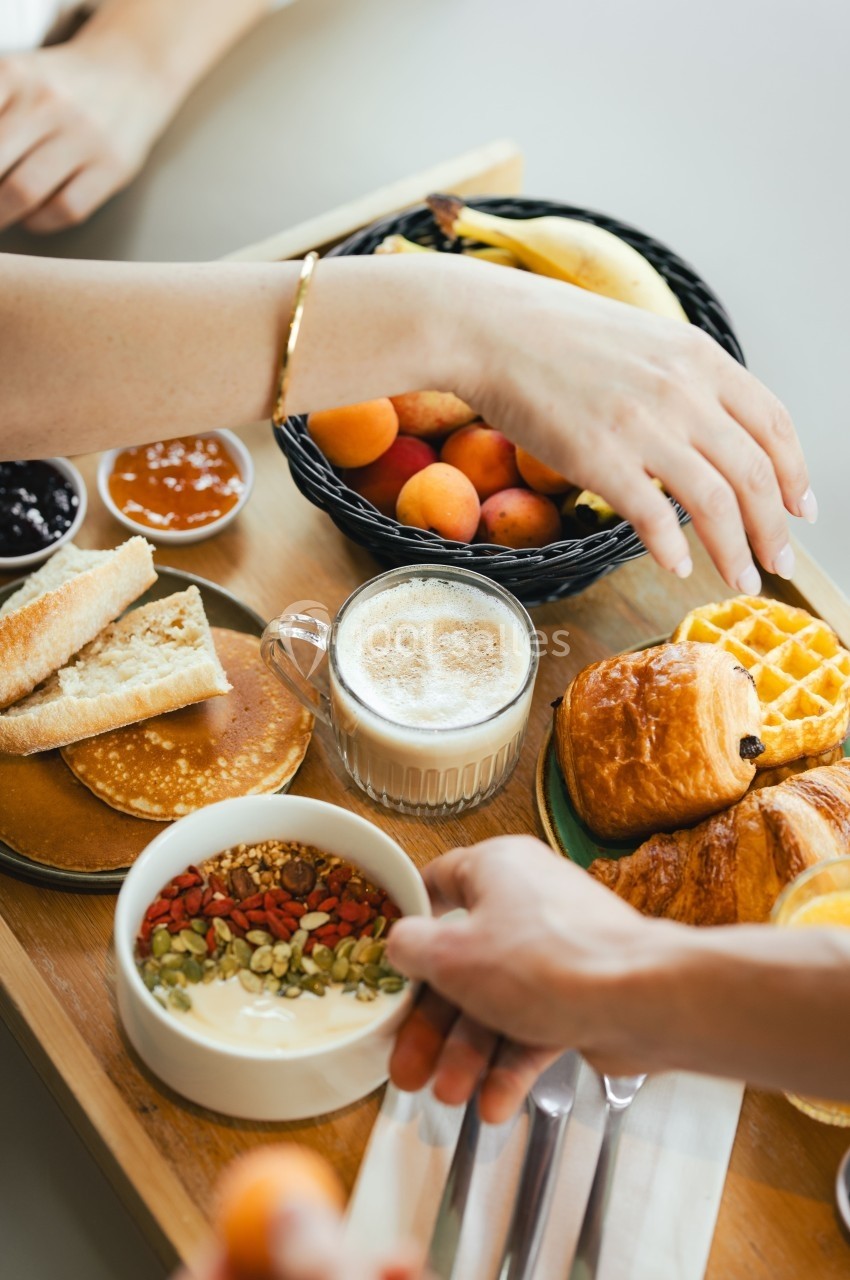 Petit-déjeuner varié avec viennoiseries, fruits, pancakes, yaourt aux graines et boisson chaude sur une table.