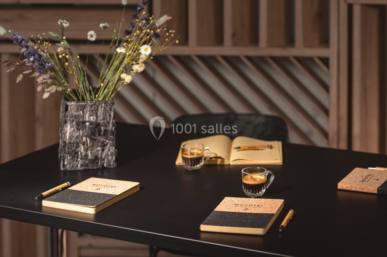 Carnet et stylos sur une table noire avec des tasses de café, un vase de fleurs séchées et un carnet ouvert en arrière-plan.