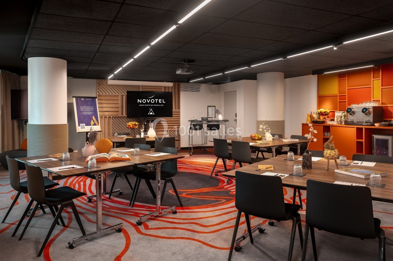 Salle de réunion moderne avec tables, chaises, écran, décorations et espace de pause avec machine à café.