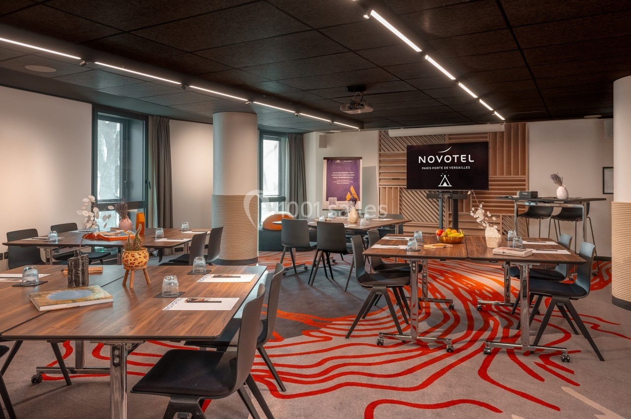 Salle de réunion moderne avec tables en bois, chaises noires, éclairage tamisé et écran affichant ’Novotel’.