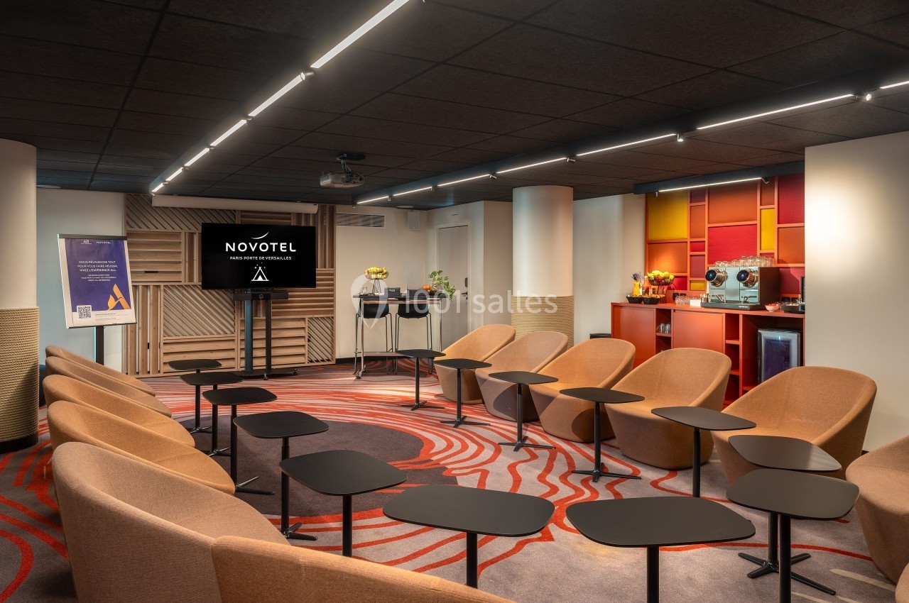 Salle de réunion moderne avec fauteuils, tables individuelles, écran de présentation et espace café en arrière-plan.