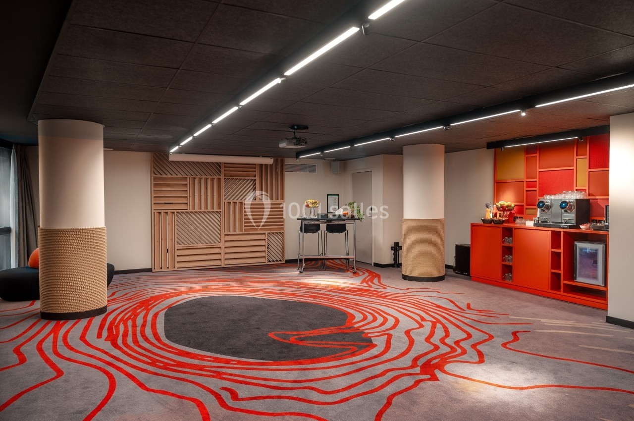Salle moderne avec tapis aux motifs rouges, colonnes blanches, éclairage au plafond et coin café équipé à droite.