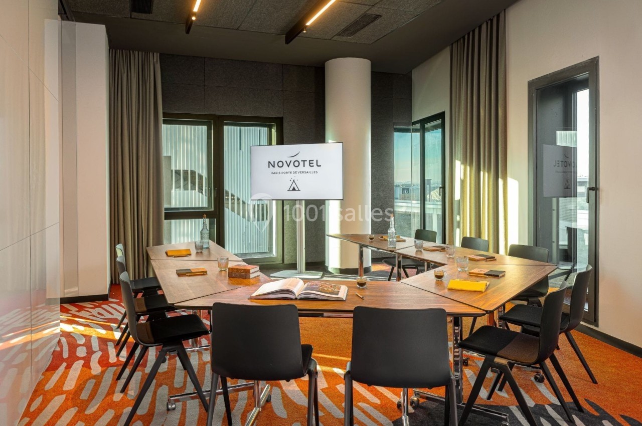 Salle de réunion lumineuse avec table en bois, chaises noires, écran affichant ’Novotel’ et grandes fenêtres vitrées.