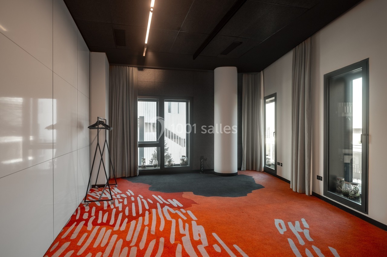 Salle moderne avec tapis rouge et noir, murs blancs et gris, grandes fenêtres et un portant à vêtements.